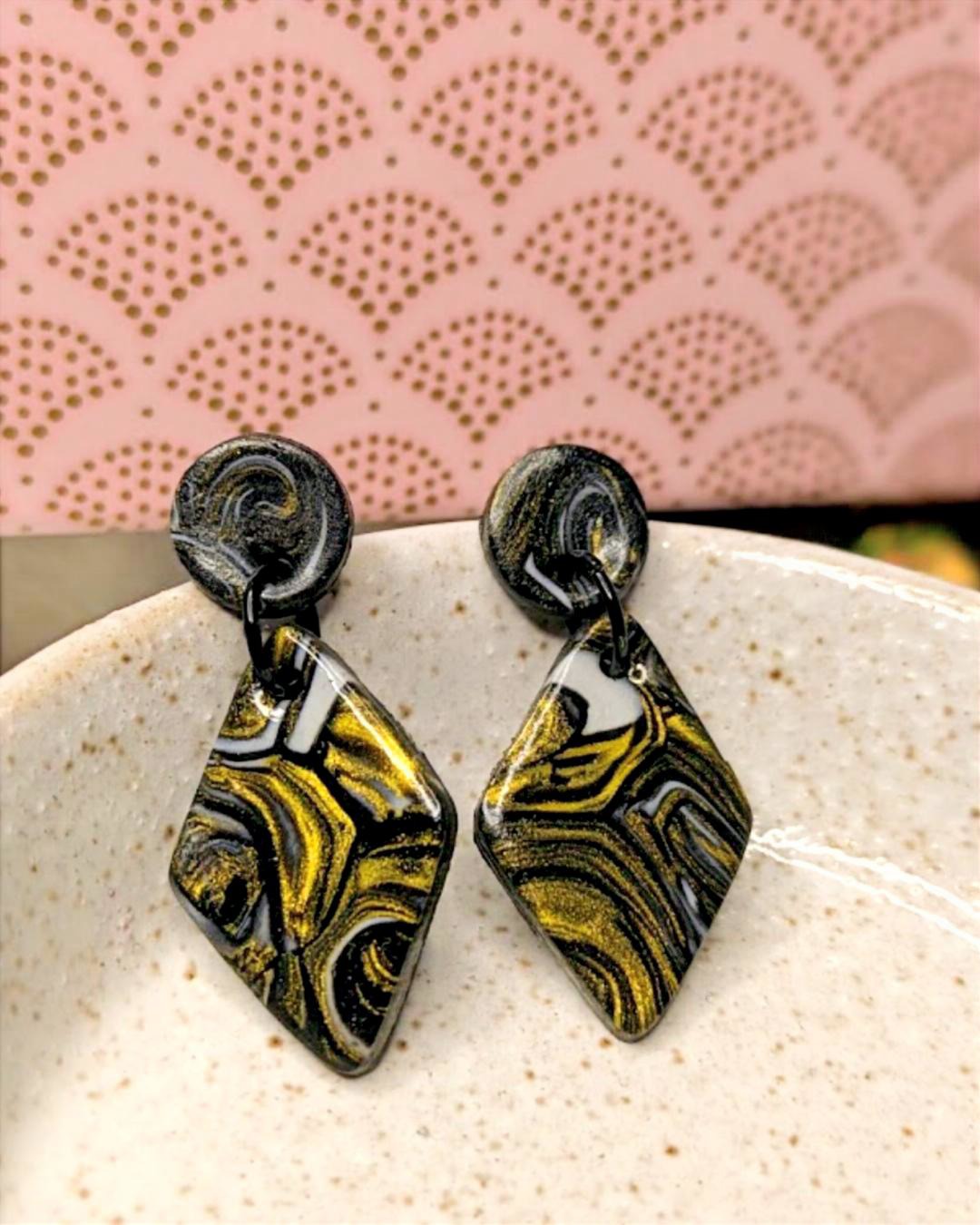 Black and gold marbled earrings on a textured surface with a pink patterned background